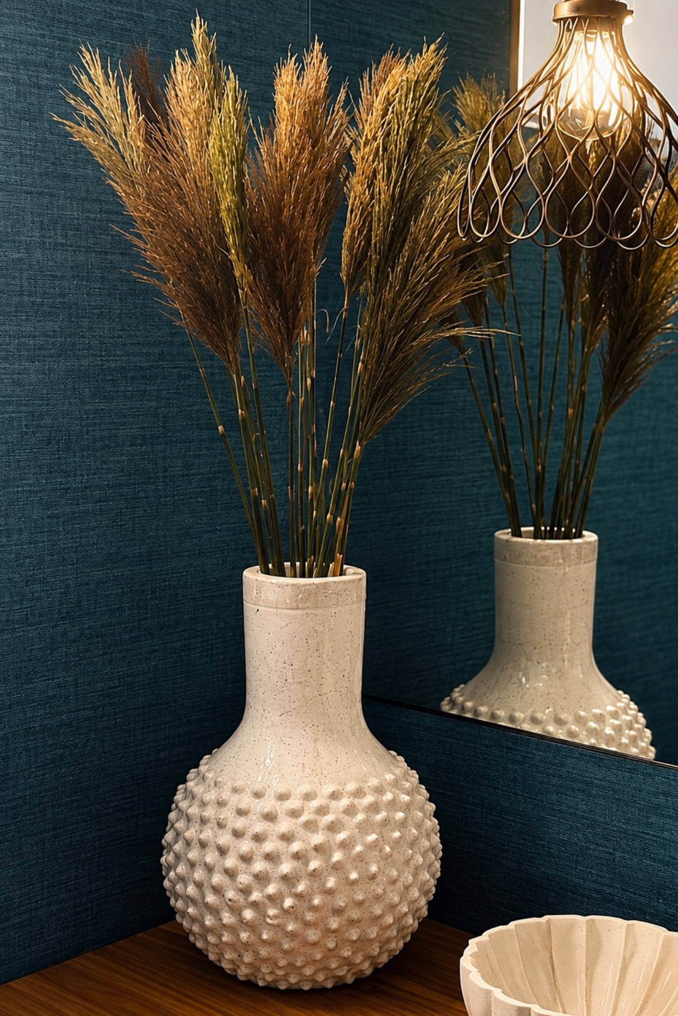 Espigas secas de pampas en un jarrón color crema con relieves frente a una pared azul petróleo
