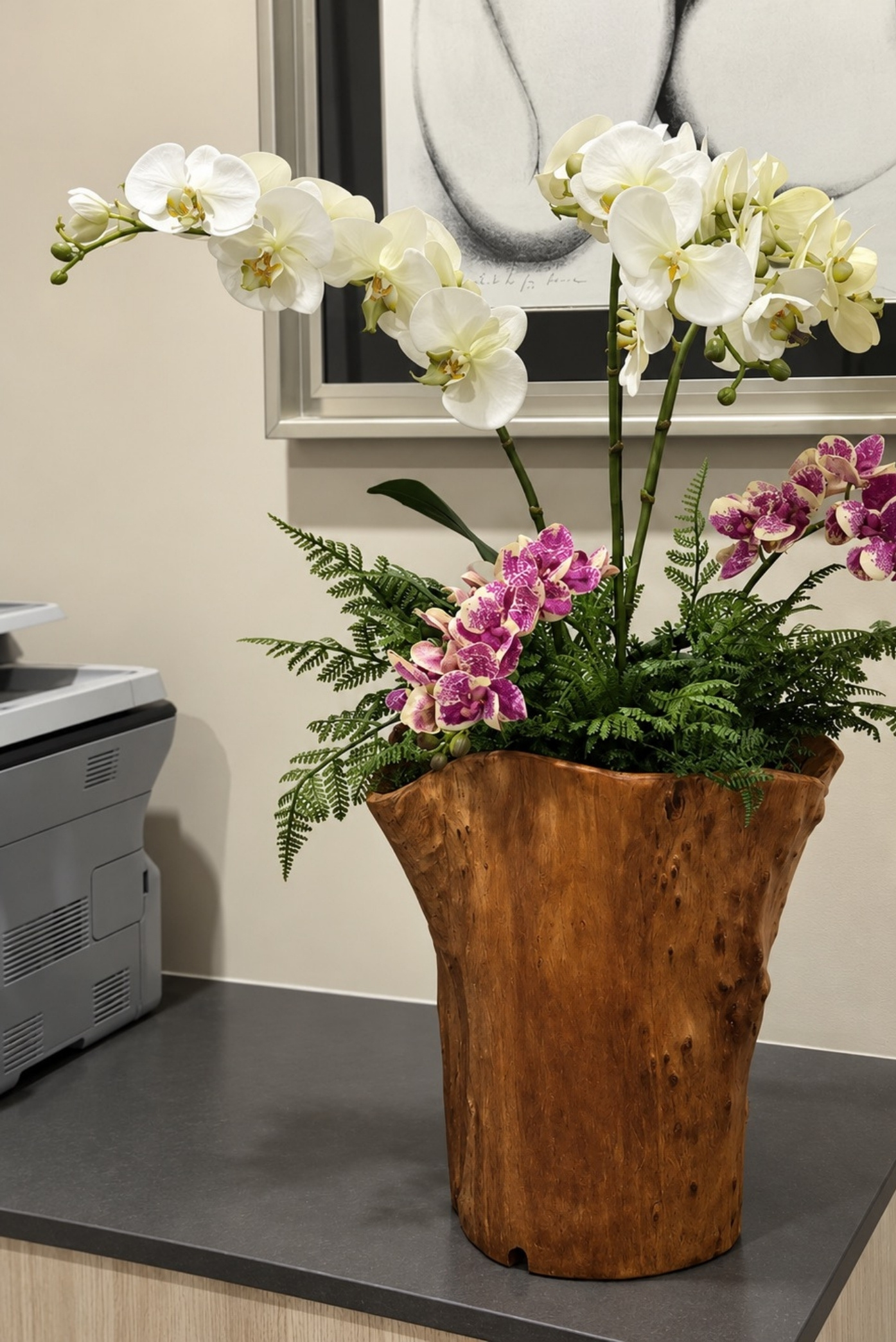 Orquídeas blancas y moradas con helechos en un jarrón de tronco de madera junto a una pared