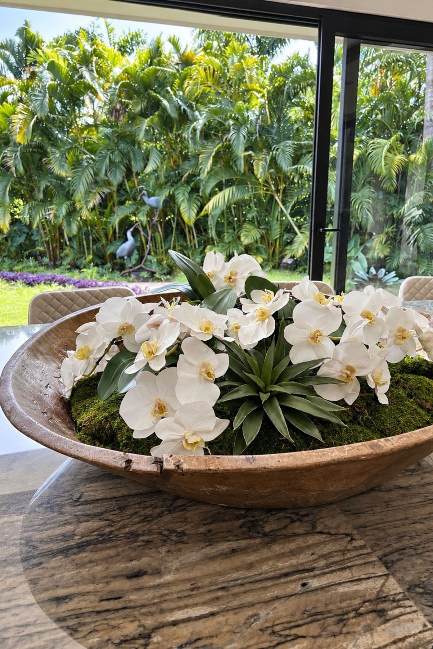 Orquídeas blancas y bromelias con musgo en un gran cuenco de madera, con un jardín tropical al fondo